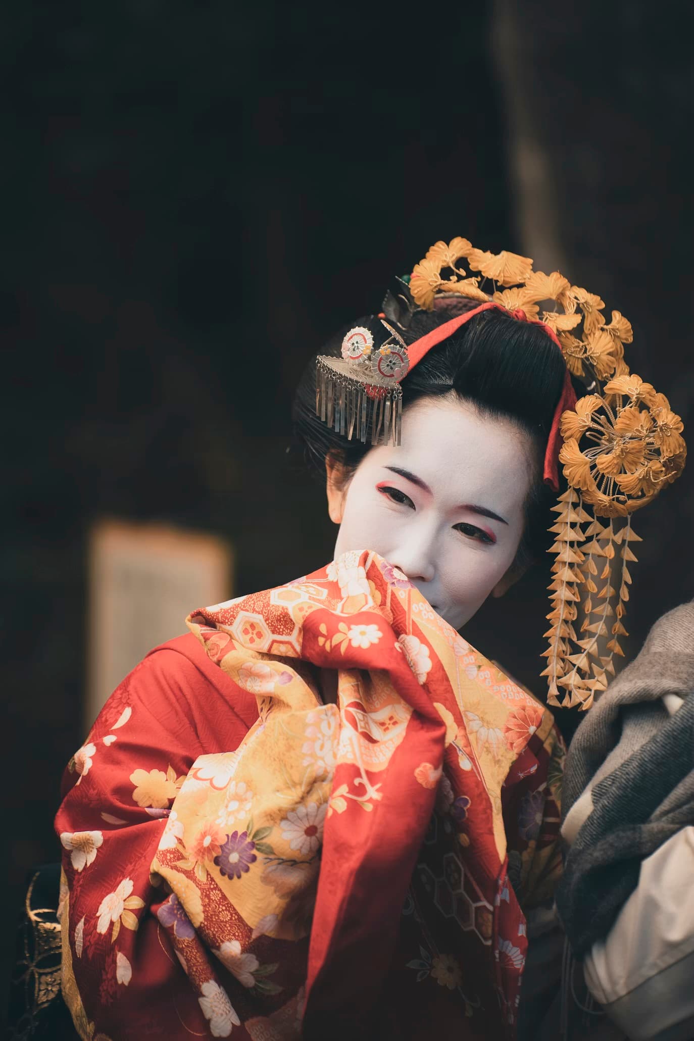 Maiko with white makeup and ornate hair ornaments partially covering her face with floral kimono.