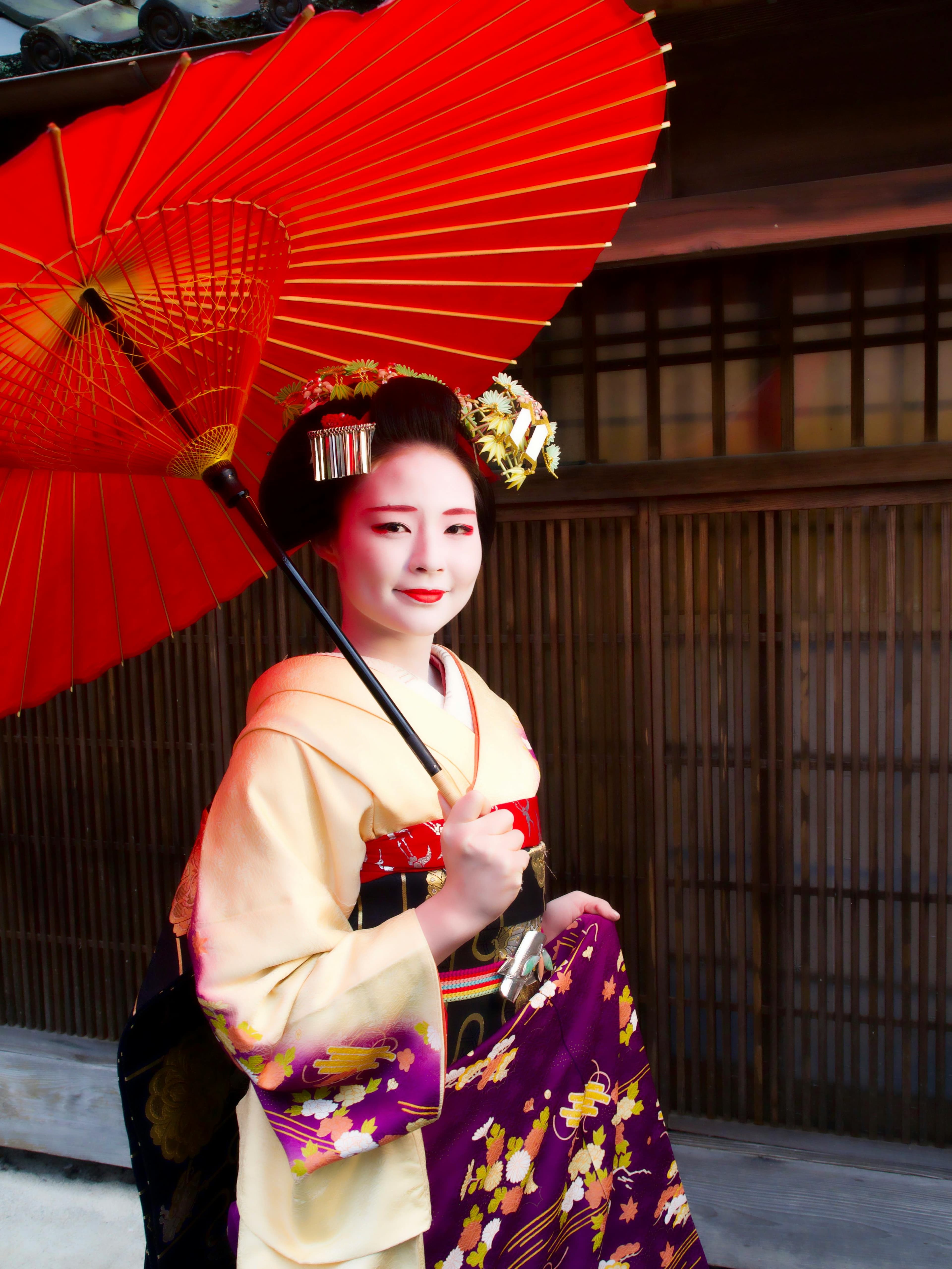 Maiko in traditional kimono and makeup holding a red wagasa umbrella against a wooden background.