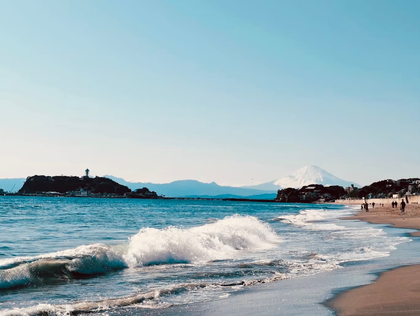 Crashing waves on a sandy beach with Mount Fuji and Enoshima Island in the distance.