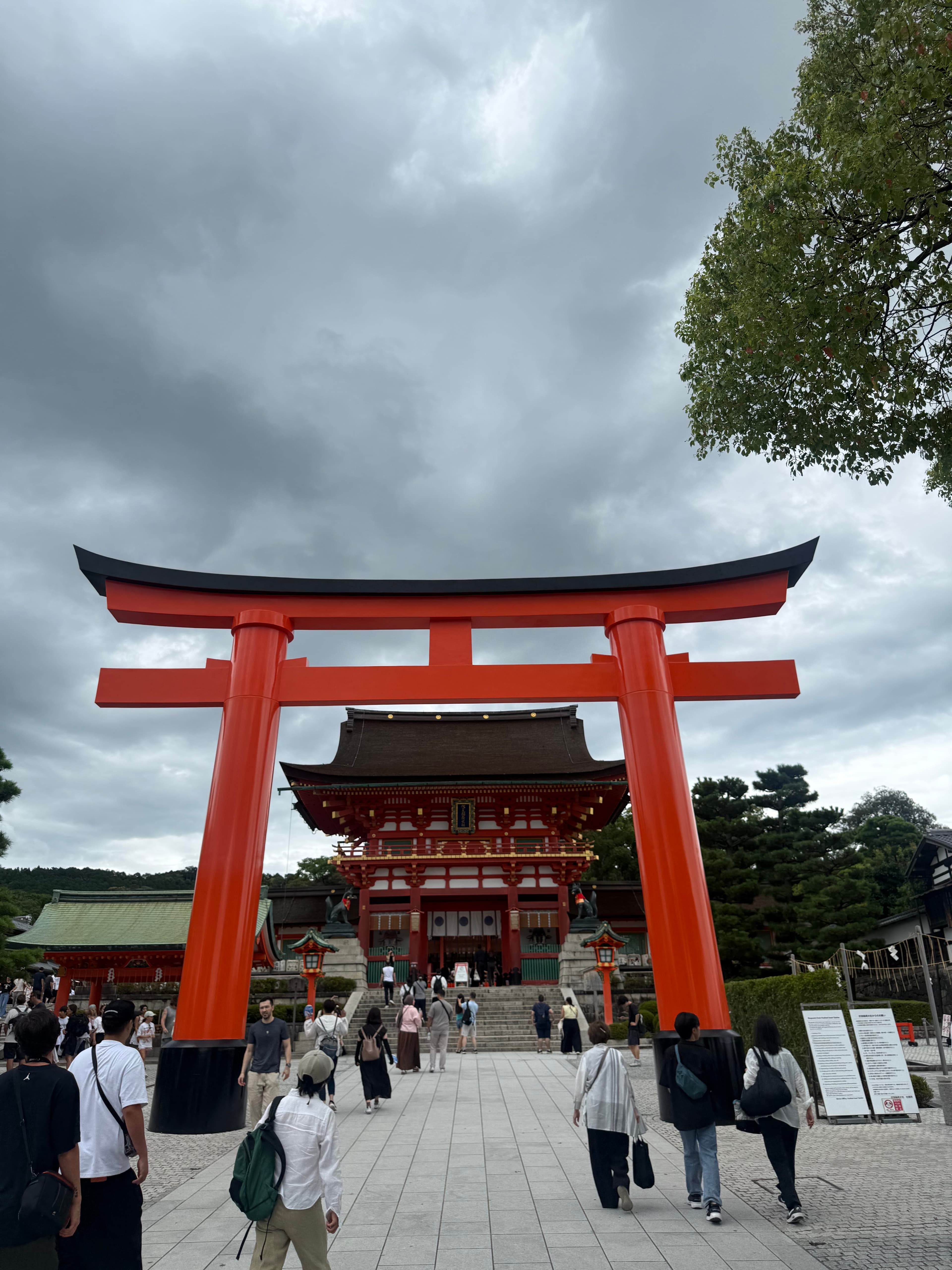 Large vermilion torii gate leading to a traditional Japanese shrine under a cloudy sky.